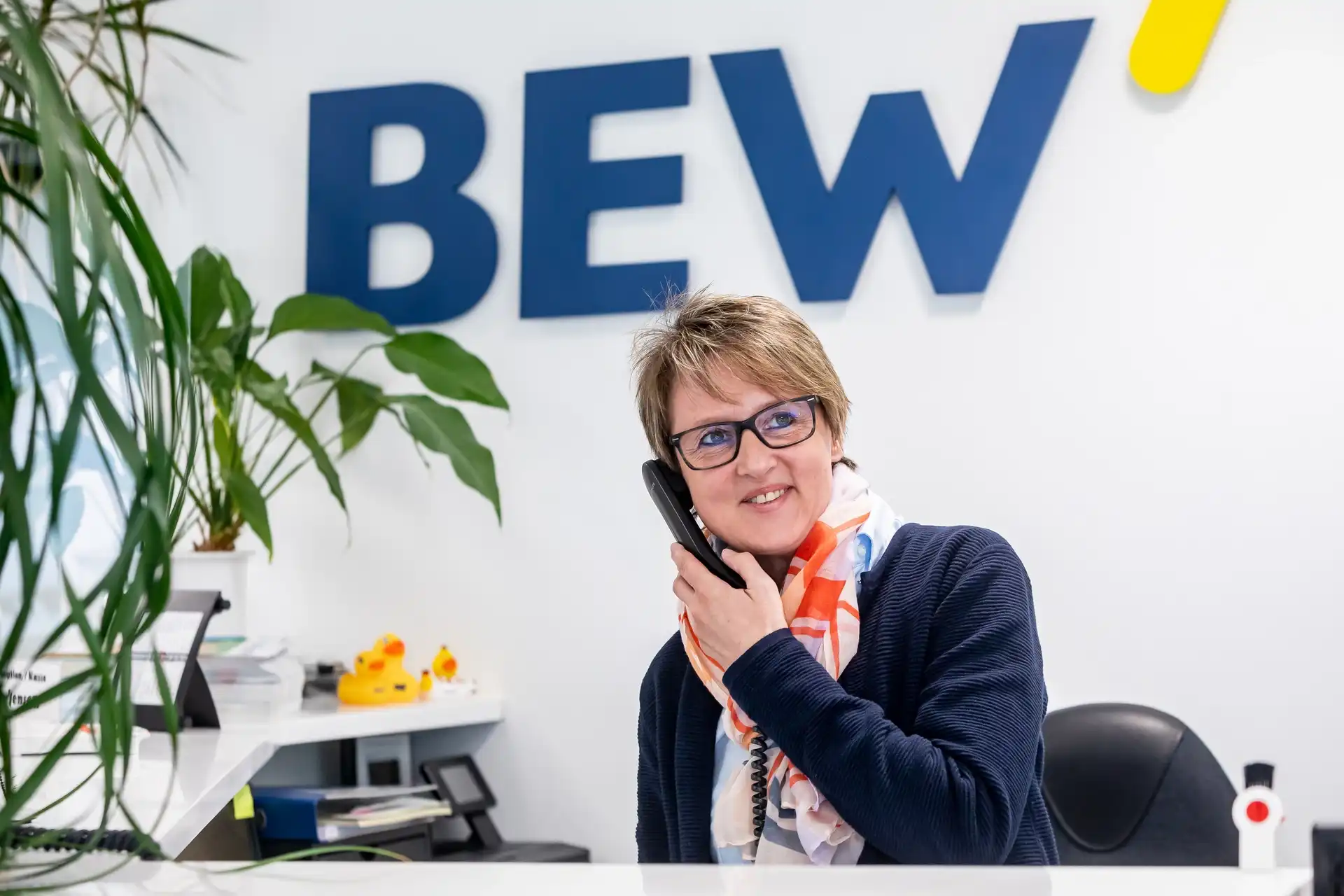 Die Mitarbeiterin Frau Jensen am Empfang, spricht am Telefon, im Hintergrund mit dem Schriftzug 'BEW' und grünen Pflanzen.